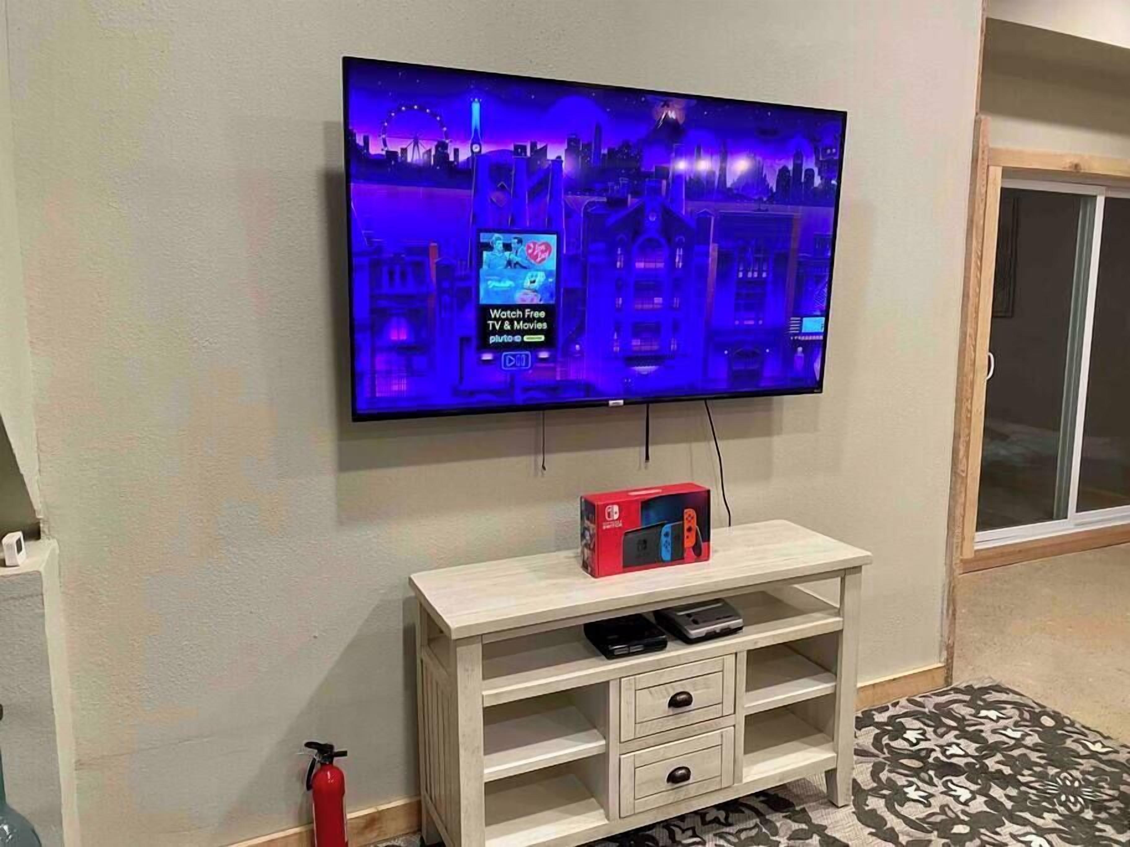 A flat-screen TV mounted on a wall, a wooden TV stand with a Nintendo Switch box, and a fire extinguisher on the floor.