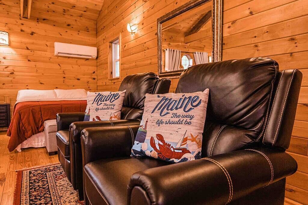 A cozy cabin room with wooden walls, a bed with red bedding, two leather chairs with 'Maine' pillows, and a large mirror on the wall.