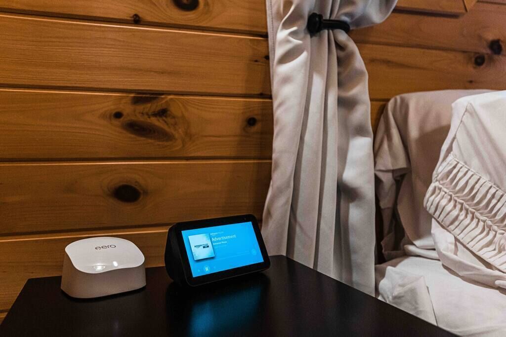 A wooden wall with a curtain, a white bed with a ruffled pillow, and a black table with a tablet and a white device.