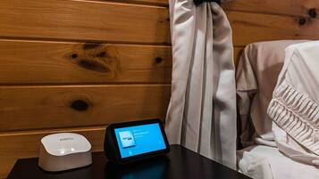 A wooden wall with a curtain, a white bed with a ruffled pillow, and a black table with a tablet and a white device.