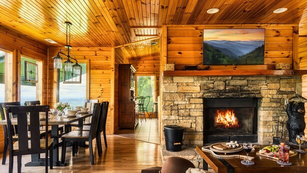 Interior acogedor de una cabaña con chimenea de piedra, comedor de madera y un cuadro grande sobre la chimenea.