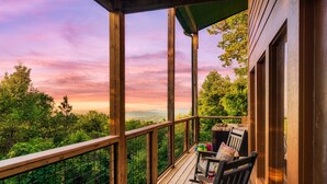 Terraza de madera con vistas a un bosque y montañas, una mesa con sillas y una zona cubierta.