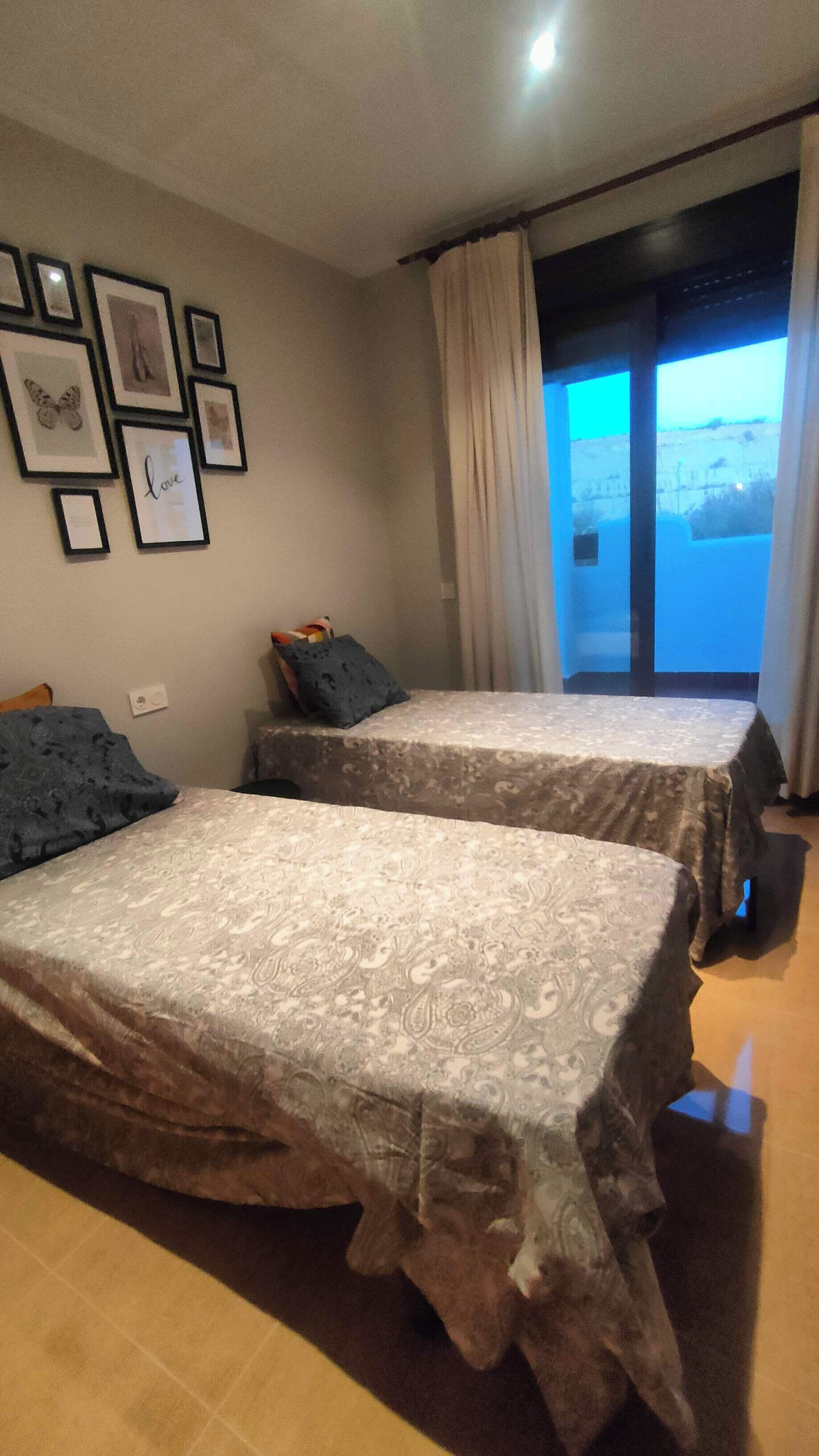 Une chambre avec deux lits, une fenêtre avec des rideaux et des tableaux encadrés accrochés au mur.