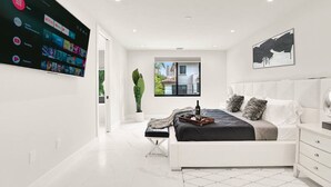 Un dormitorio moderno con un televisor de pantalla plana grande mostrando la interfaz de un servicio de streaming, una cama con ropa de cama en tonos grises y negros, una mesita de noche y una ventana con vistas a edificios.