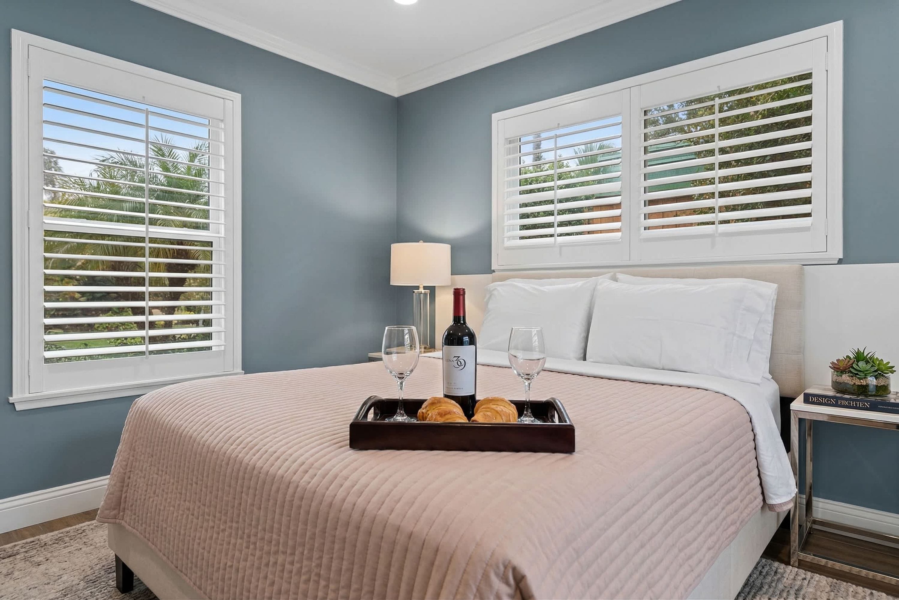 A bedroom with a bed, a tray with wine and pastries, a lamp, and a book on a nightstand.