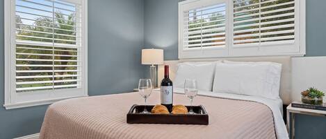 Une chambre avec un lit, un plateau avec du vin et des pâtisseries, une lampe et un livre sur une table de chevet.