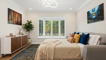 Une chambre à coucher avec un lit, une fenêtre avec des volets, une plante, une armoire et des œuvres d’art accrochées aux murs.