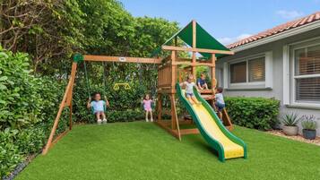 A wooden play structure with a slide and swings, surrounded by well-maintained grass and greenery.