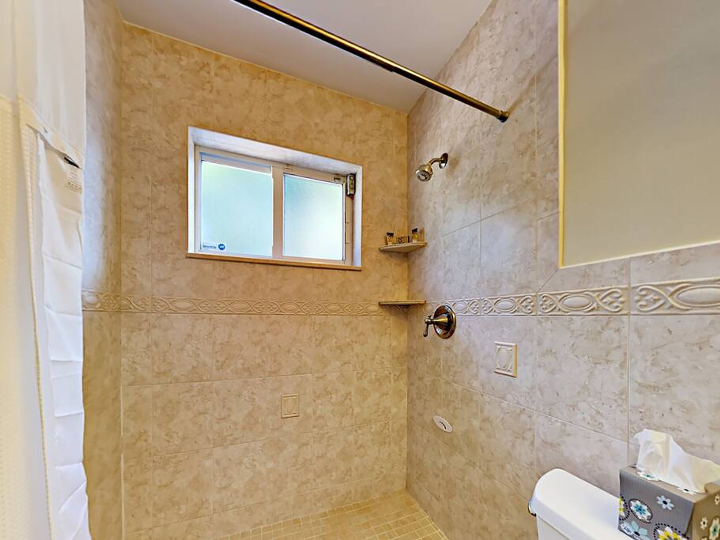 A bathroom with beige tiles, a window, a shower, a toilet, and a towel rack.