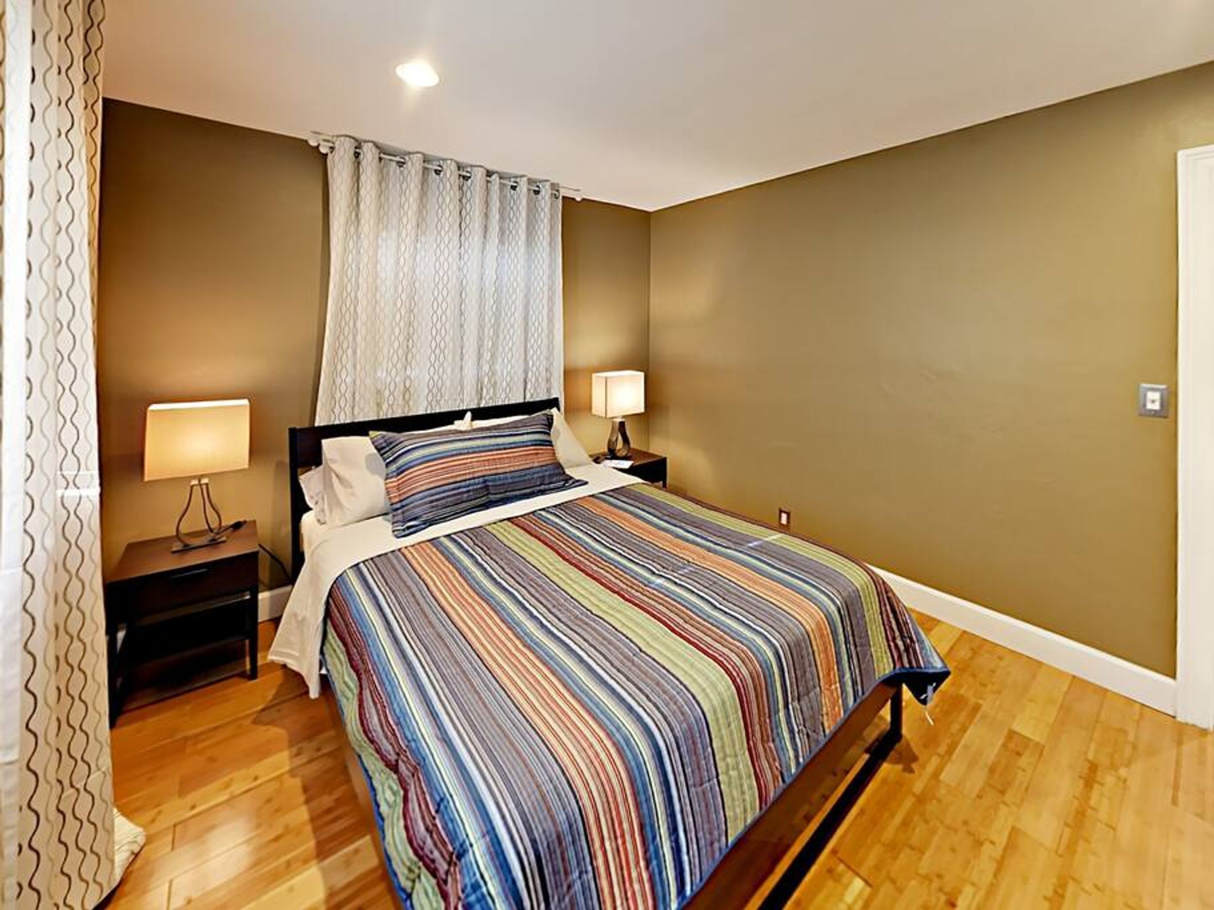 A bedroom with a bed, two bedside tables, a window with curtains, and a wooden floor.