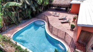 Una piscina de borde curvo, rodeada por un patio de ladrillo y una estructura con techo de paja.