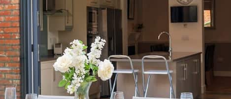 Un coin repas avec une table dressée pour quatre personnes, un vase de fleurs et une cuisine en arrière-plan.