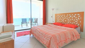 A hotel room with a bed, a nightstand, a chair, a table, and a view of the sea through a sliding glass door. - Windhouse  (Imsouane)