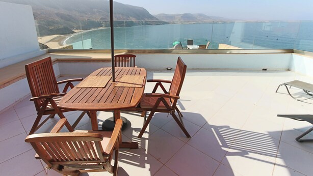 A wooden table and chairs with a red umbrella on a balcony overlooking a beach and ocean. - Windhouse  (Imsouane)