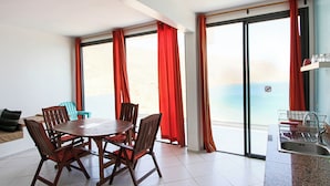 A room with a dining table and chairs, a kitchen area with a sink, and large windows with red curtains. - Windhouse  (Imsouane)