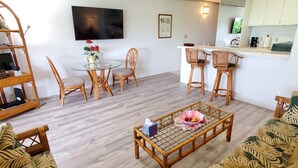 A living room with a glass table, wicker chairs, a sofa, a coffee table with fruit, a TV, and a kitchen area with white cabinets. - Condo in the center of Kona, 2-unit AC, Washer/Dryer, Full Kitchen, Free Parking (Kailua-Kona)
