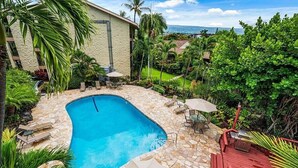 A swimming pool with stone tiles, surrounded by lounge chairs and umbrellas, with a view of a building and greenery. - Condo in the center of Kona, 2-unit AC, Washer/Dryer, Full Kitchen, Free Parking (Kailua-Kona)