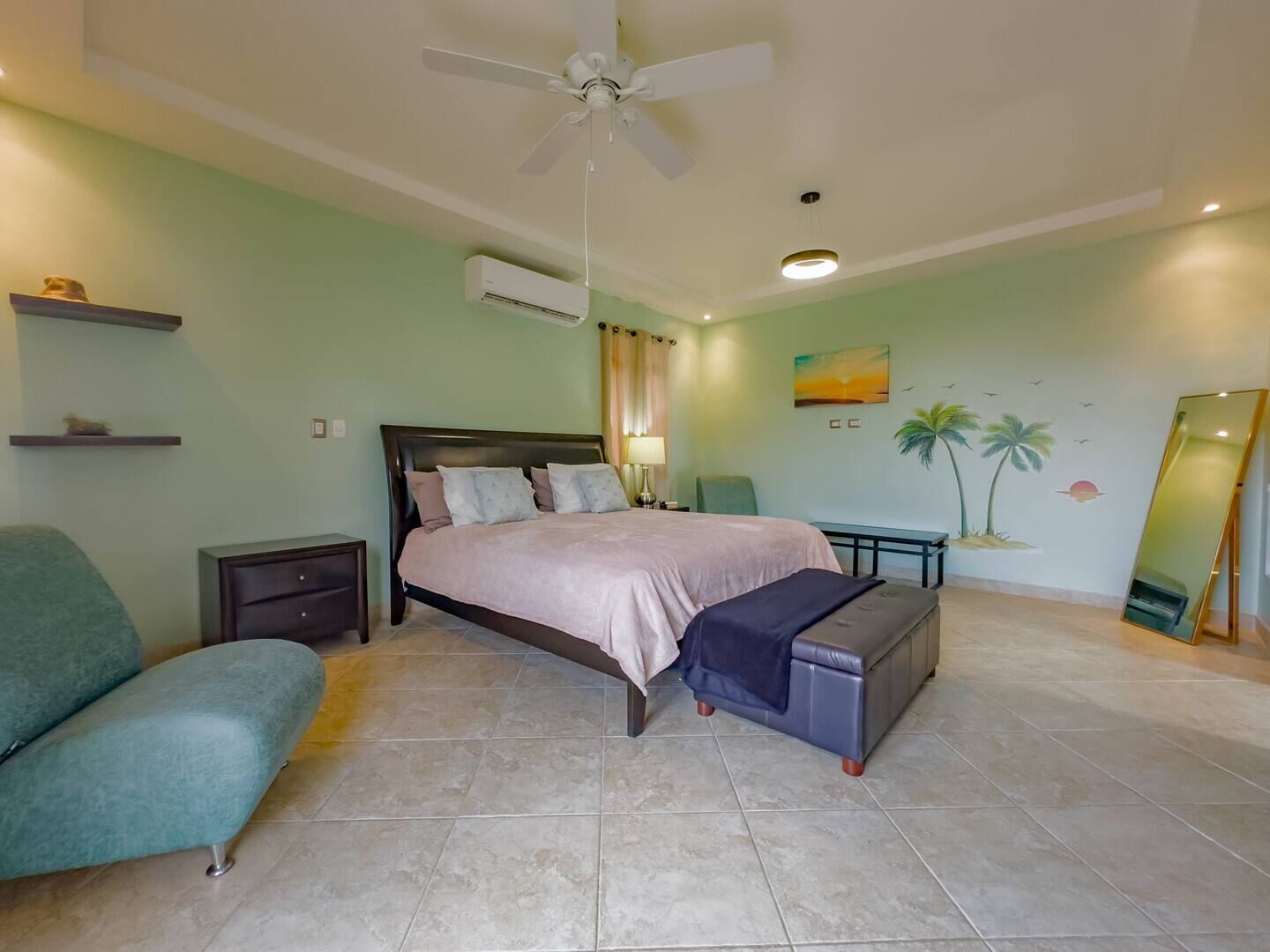 A bedroom with a bed, a nightstand, a bench, a chair, a ceiling fan, an air conditioning unit, a wall-mounted mirror, and a wall with a palm tree mural.