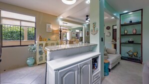Una cocina amplia con una isla central, un comedor con sillas de mimbre y una sala de estar con sofá.