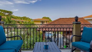 Un balcón con dos sillas azules, una mesa negra y vistas a casas con techos de tejas rojas.