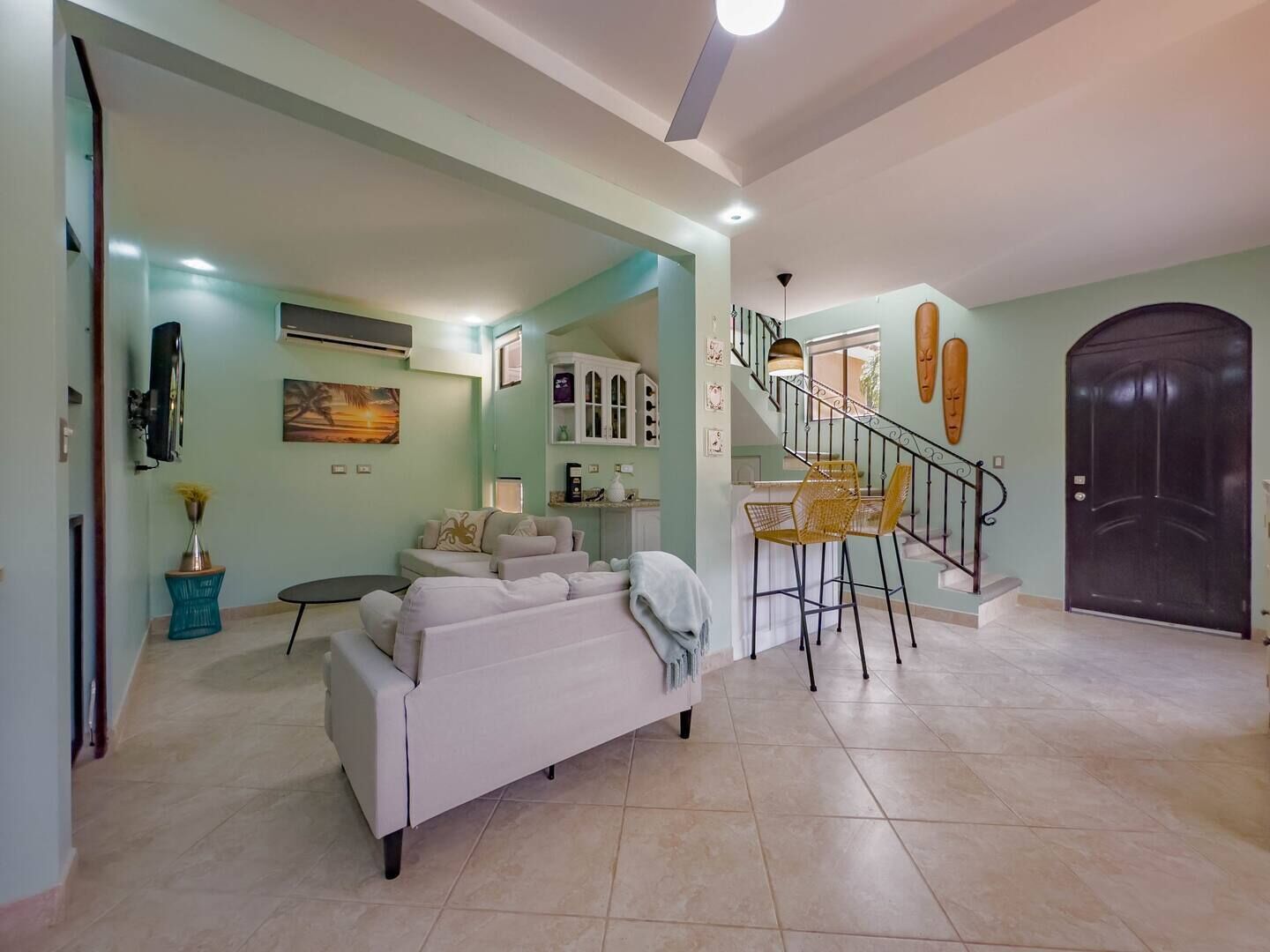 A spacious living area with a sofa, a dining table with chairs, a staircase, and a dark door.