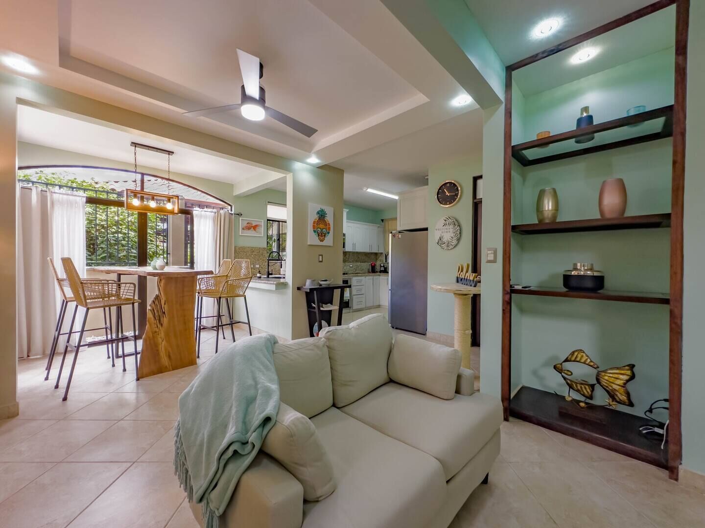 A modern living room with a sofa, a dining area with chairs, a kitchen with cabinets, and a ceiling fan.