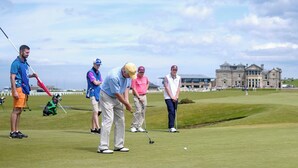 Un terrain de golf avec des joueurs et des spectateurs, un bâtiment historique en arrière-plan et un ciel dégagé.