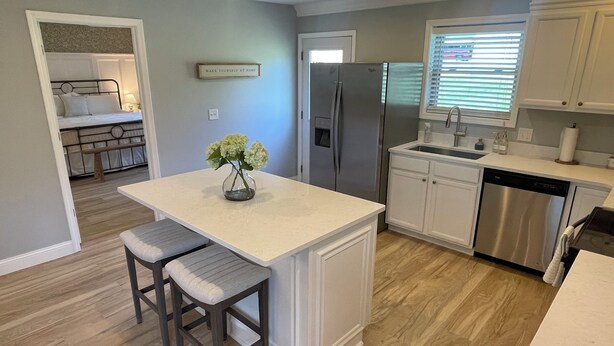 A modern kitchen with white cabinetry, a stainless steel refrigerator, and a built-in dishwasher. There is a small island with two stools and a vase of flowers. - Laura’s Simply Charming Home on Harrison (Quincy)
