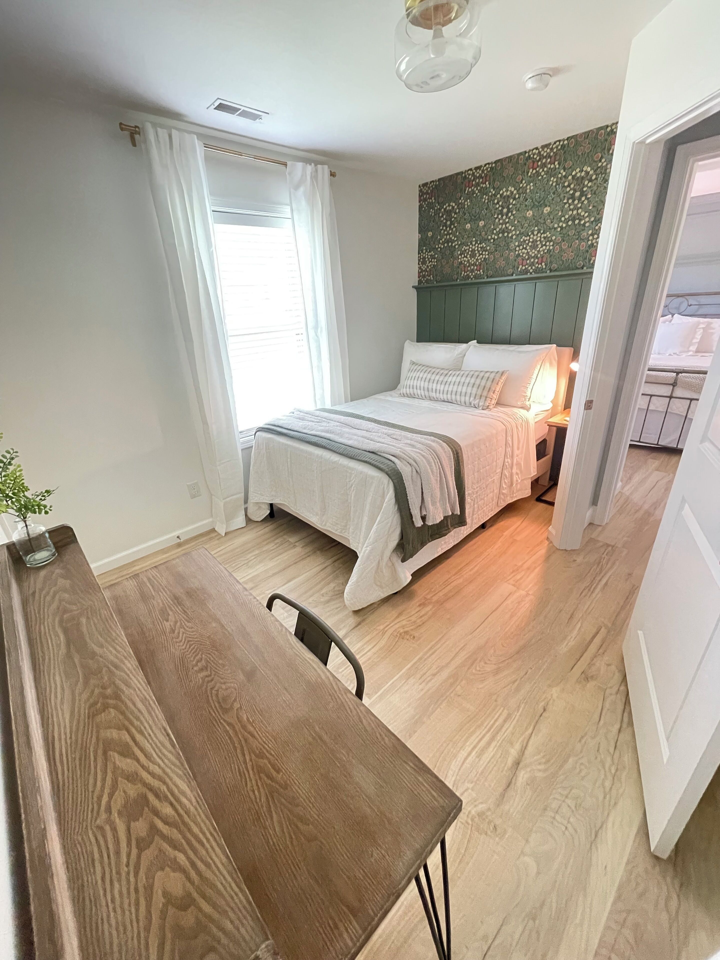 Een slaapkamer met een houten bed, een nachtkastje, een stoel, een raam met witte gordijnen en een groene, behangen muur.