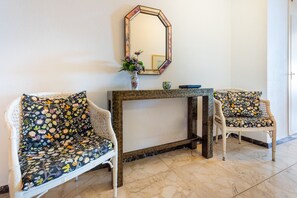 Una habitación con dos sillas blancas de mimbre con tapicería floral, una mesita con un florero y un espejo en la pared.