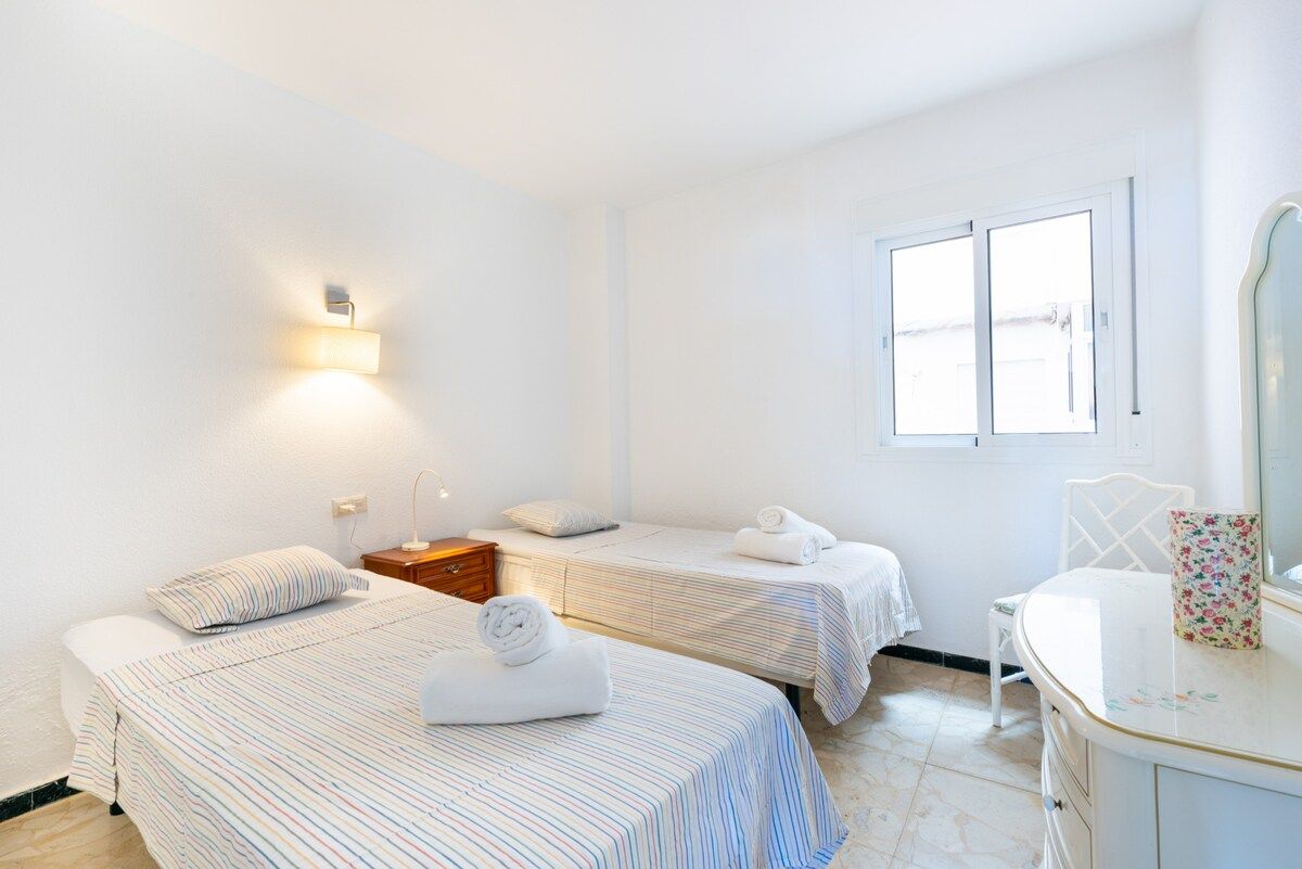 Ein Hotelzimmer mit zwei Einzelbetten, einem kleinen Nachttisch, einem Stuhl, einem Spiegel und einem Fenster.