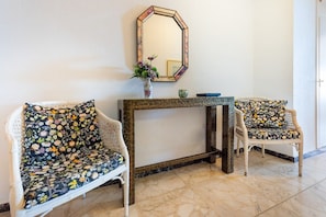 A room with two white wicker chairs with floral upholstery, a marble-tiled floor, and a wall-mounted mirror above a console table with a vase of flowers. - Maritimo Rey Fuengirola | Charming Beachfront Apartment with Sea View Balcony (Fuengirola)