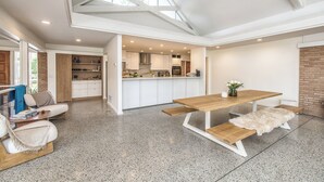 Un'ampia zona giorno con un tavolo da pranzo in legno, un divano e una panca. La stanza ha un soffitto alto con lucernari e una cucina sullo sfondo.