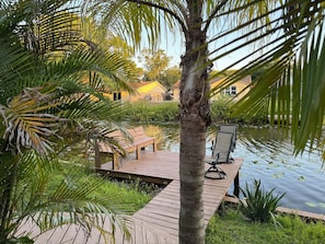 Une terrasse en bois avec un banc et une chaise, donnant sur une étendue d’eau calme parsemée de nénuphars, entourée d’une végétation luxuriante et avec un bâtiment jaune en arrière-plan.