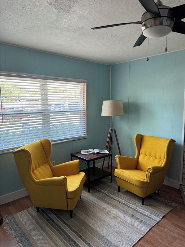 Une pièce avec deux fauteuils jaunes, une petite table sur laquelle se trouve un livre, un lampadaire et un ventilateur de plafond.
