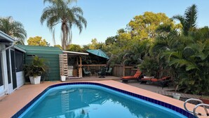 Ein Schwimmbecken mit einer Fliesenverkleidung, umgeben von einer Sonnenterrasse mit Liegestühlen und Grillplatz.