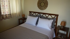 Een netjes opgemaakt bed met wit en grijs beddengoed, twee nachtkastjes met lampen, een rond decoratief object aan de muur en een raam met gordijnen.