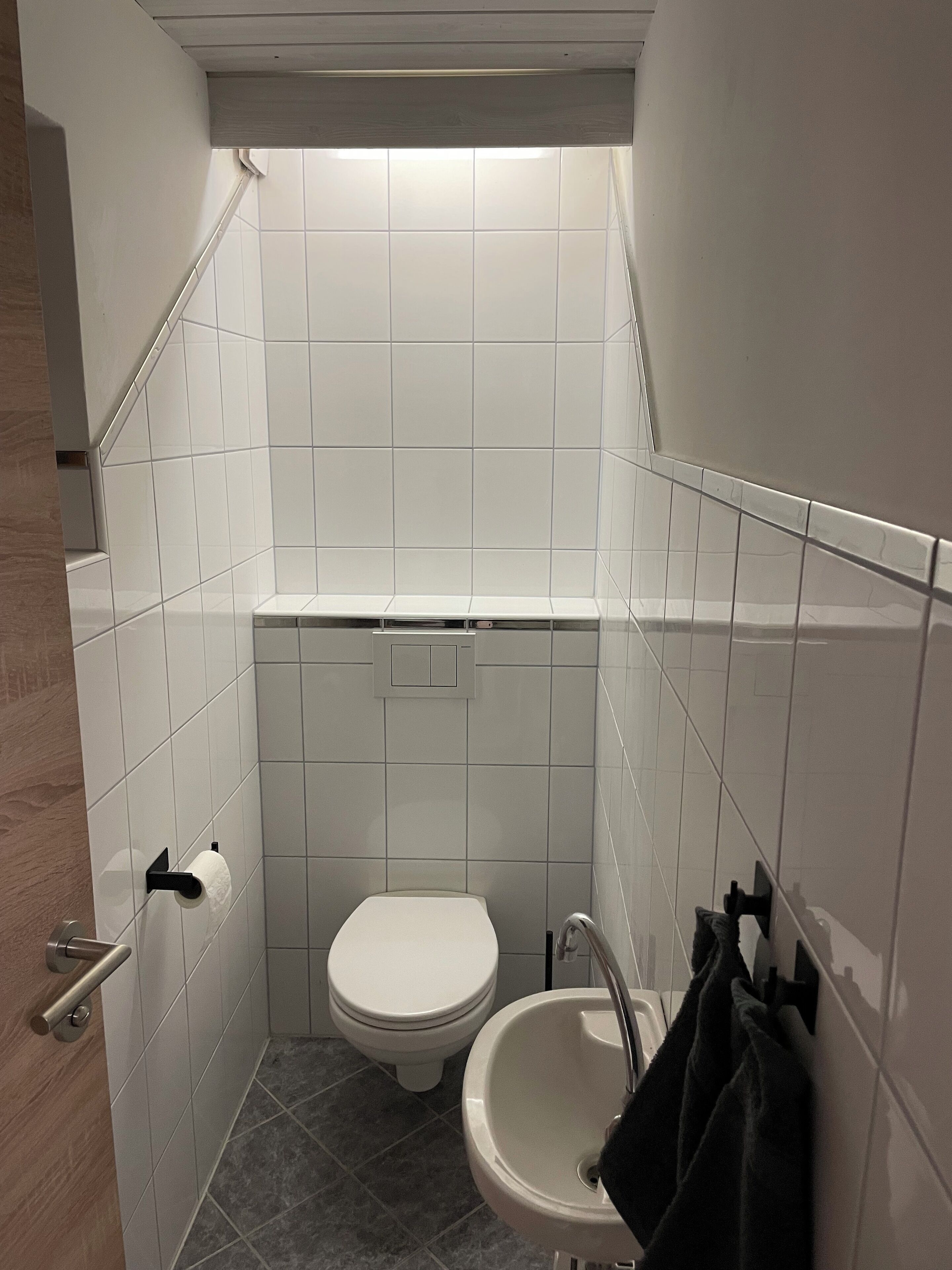 A small bathroom with white tiles, a toilet, a sink, and a towel rack.