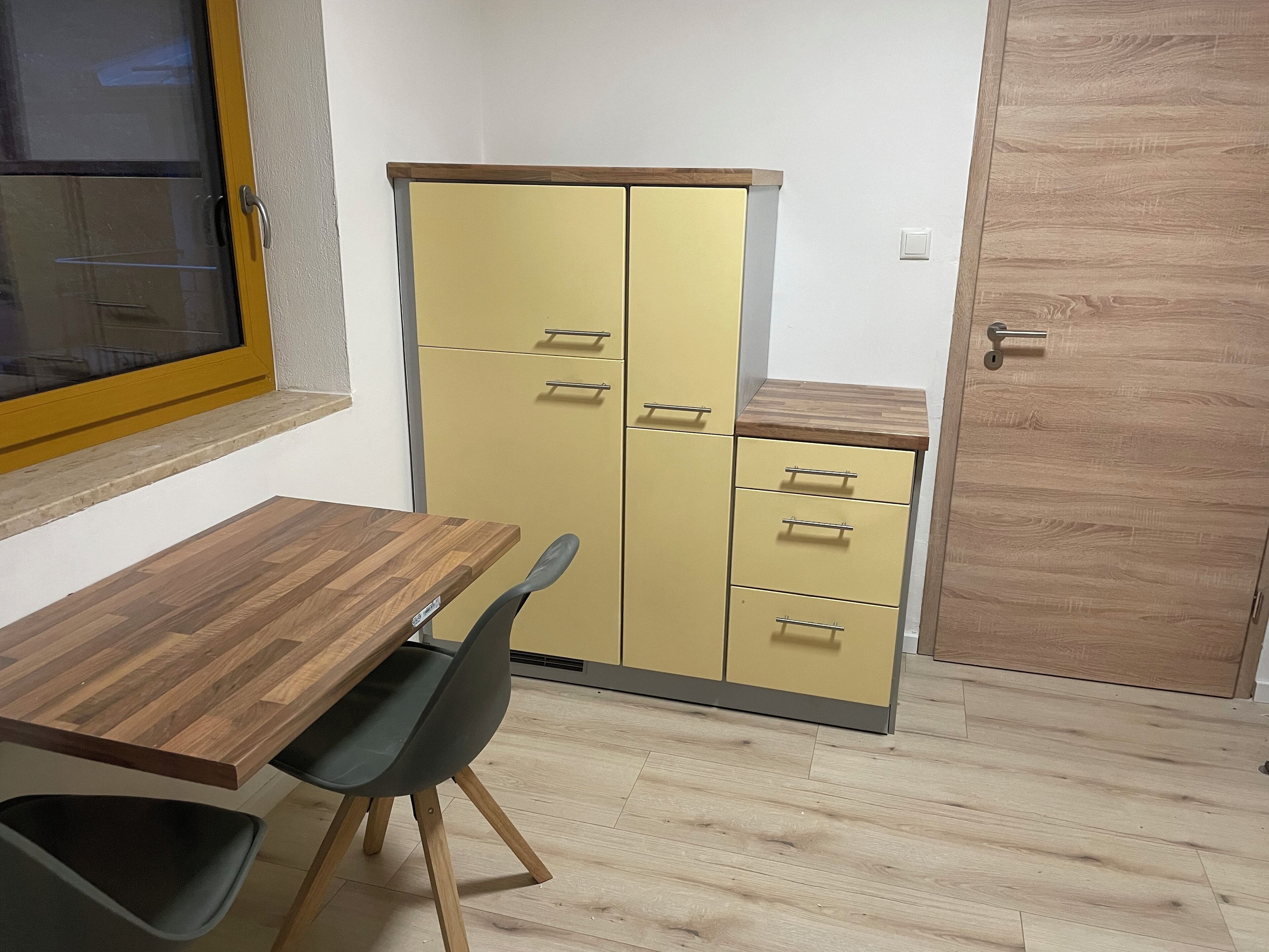 Una habitación pequeña con una mesa de madera, una silla negra, un armario amarillo con cajones y una puerta de madera.
