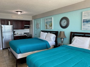 A hotel room with two beds, a kitchenette, a clock, and artwork on the walls.
