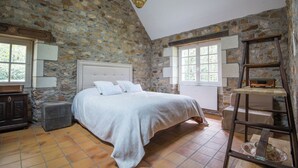 Quarto com parede de pedra, cama com roupa de cama branca, duas luminárias de cabeceira, cômoda de madeira e escada com rádio.