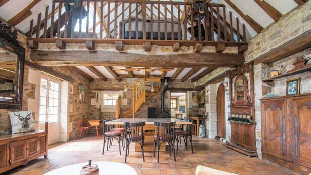 Interior rústico com parede de pedra, vigas de madeira, uma mesa central com cadeiras e uma escadaria que leva a um andar superior.