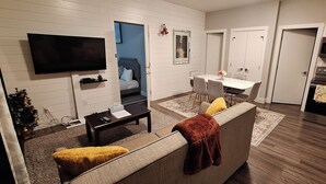 En moderne stue med et fladskærms-TV, et spiseområde med bord og stole, et køkken med rustfrie hvidevarer og et lille juletræ.