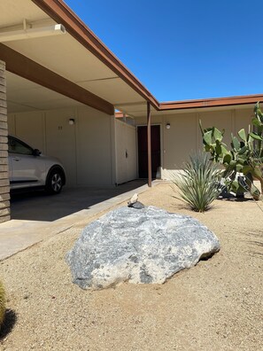 Un garaje con un coche aparcado, una roca grande y plantas del desierto.