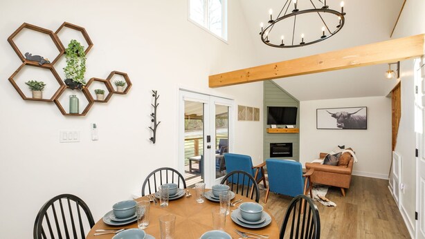 A dining area with a round wooden table set for six, surrounded by black chairs. A hexagonal wooden shelf with plants and decorative items is mounted on the wall above the table. - Private, Hot tub, Firepit, Bocce Ball, Horse Shoes, Outdoor Ping Pong Table (Shenandoah)
