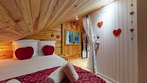 Une chambre confortable dans un chalet en bois, avec un lit, des oreillers en forme de cœur rouge, une fenêtre donnant sur la forêt et des décorations en forme de cœur sur le mur.
