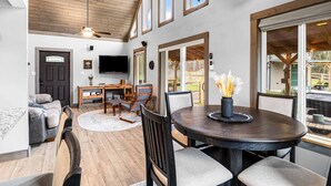 Un vaste espace de vie doté d’un plafond en bois, de grandes fenêtres, d’une table à manger ronde et d’un coin salon confortable.