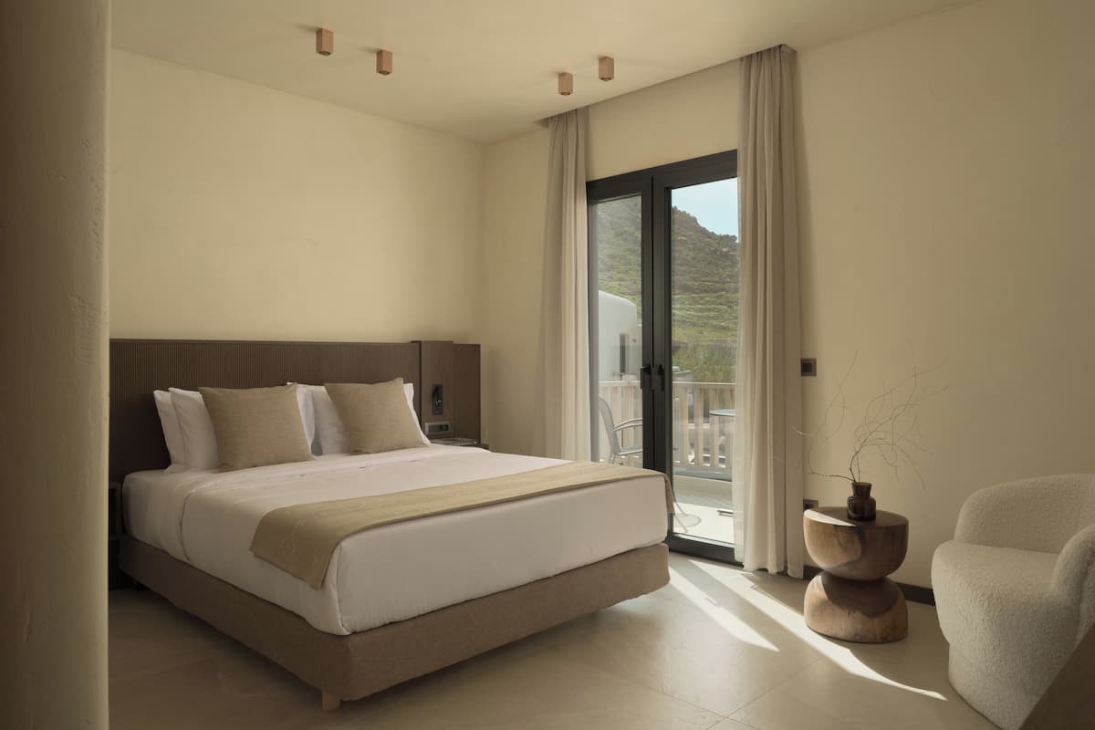 A modern hotel room with a large bed, a balcony with a view of the hills, and a small side table with a vase.