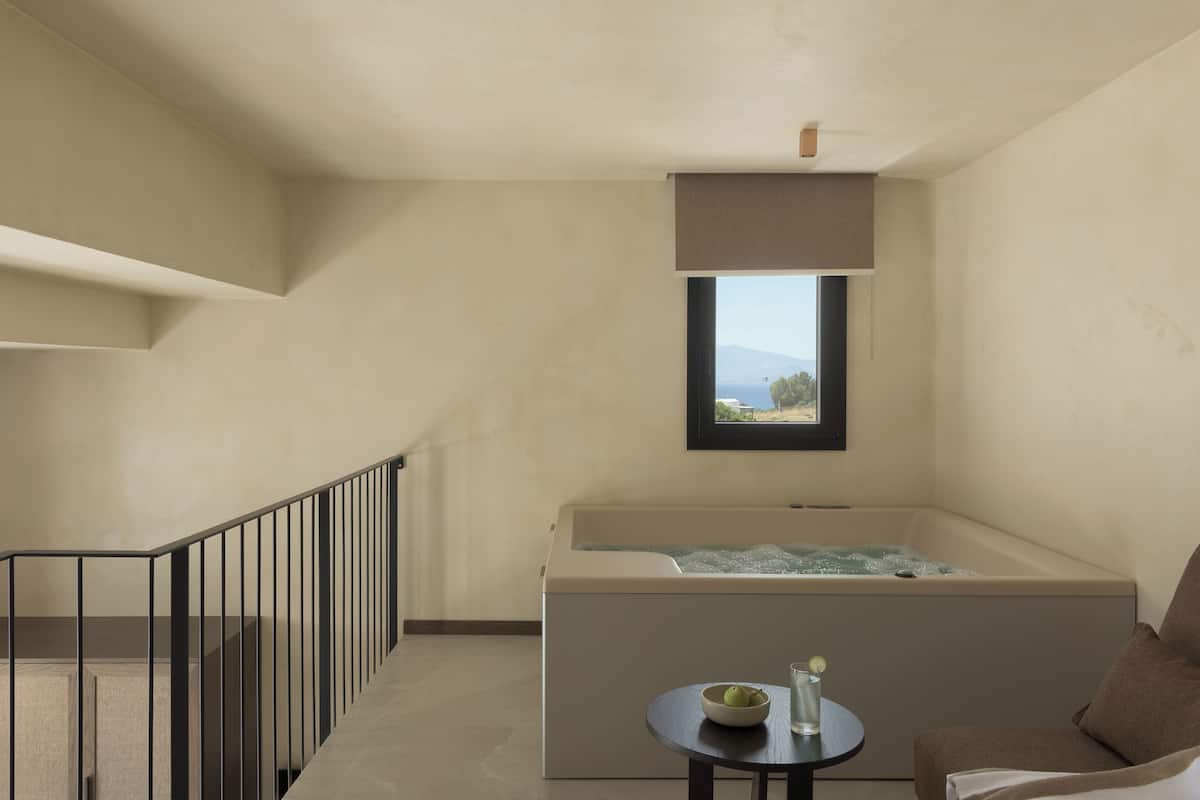 A modern room with a bathtub, a small table with a bowl and a glass, and a view of the sea through a window.