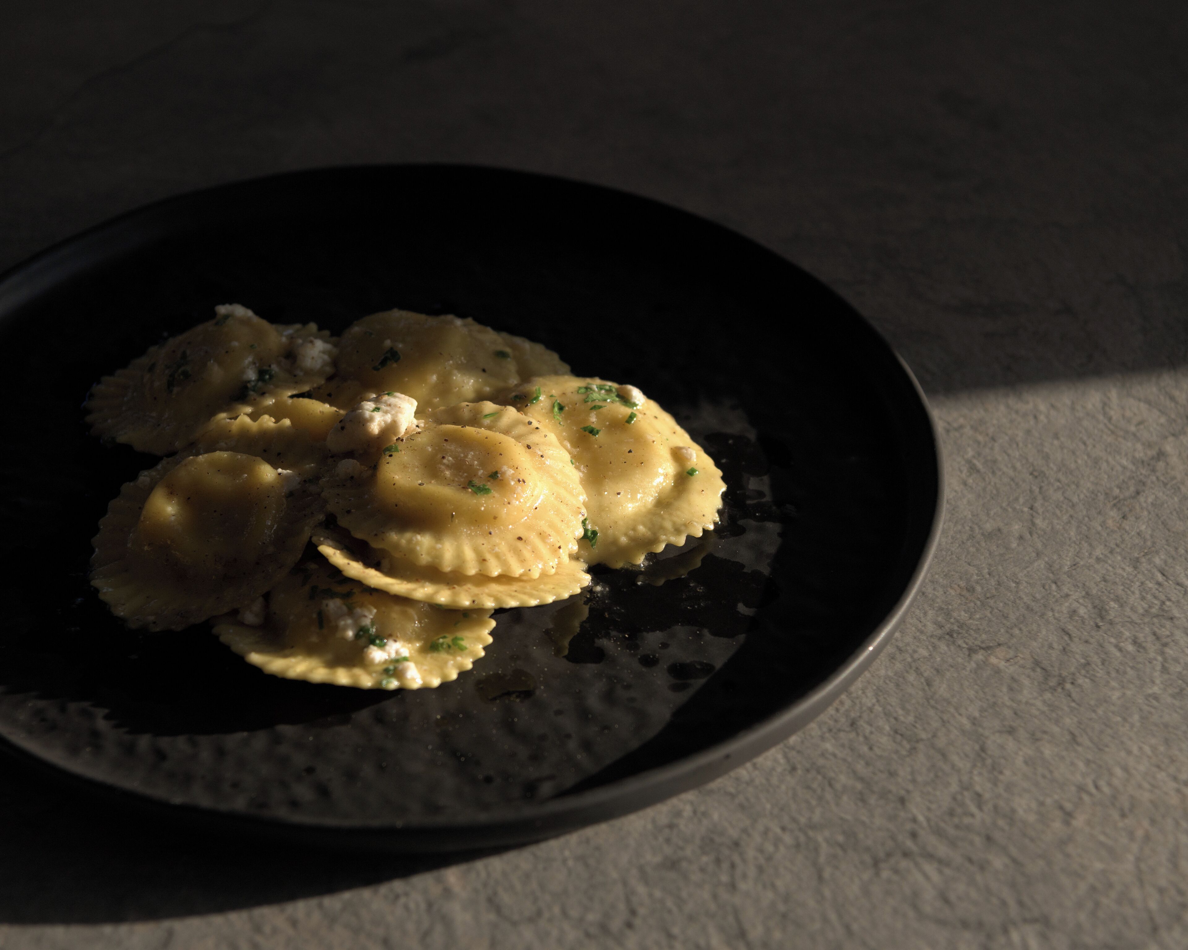 A black plate with ravioli and a small amount of sauce, placed on a textured surface.